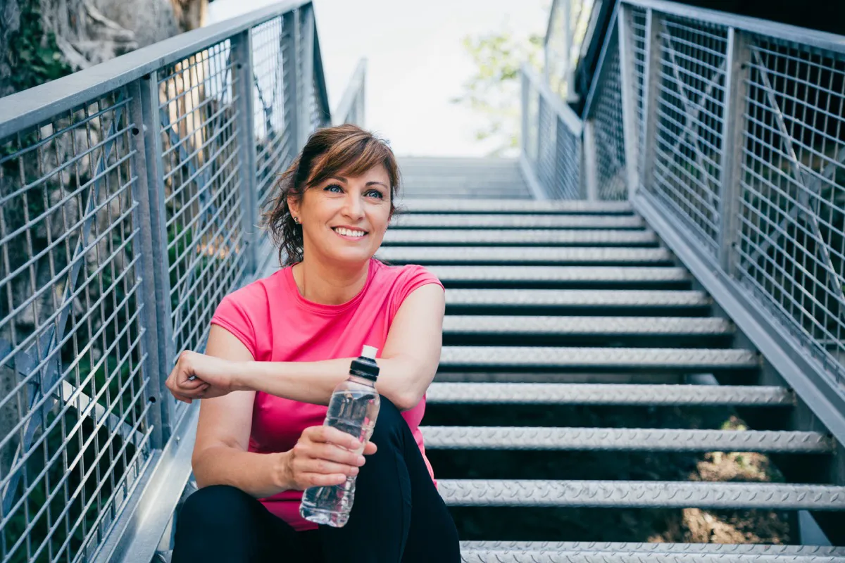 middle aged woman sitting on metallic stairs relaxing before running outdoors holding a water bottle