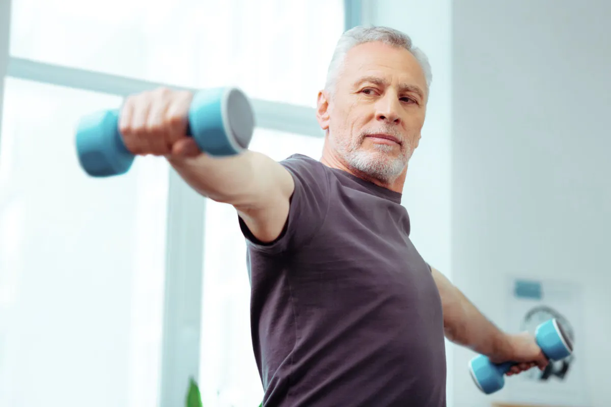 Strong fit aged man looking at his hand while training with dumbbells