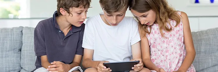 Three children sitting on a sofa watching a tablet that is in the hands of the child in the middle
